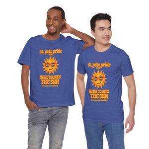 May include: Two people wearing blue t-shirts with the text "St. Pete Pride 2020" and a smiling sun graphic with rainbow-colored sunglasses. The shirts also read "Here Comes The Sun". The people are wearing blue jeans.