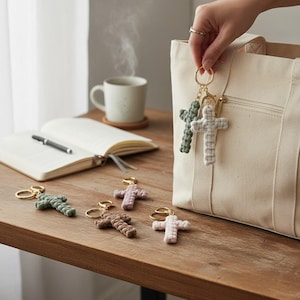 May include: Macrame cross keychains in various colors, including green, brown, and pink, with gold-colored key rings. A hand holds two keychains near a canvas tote bag. Other keychains are arranged on a wooden surface, alongside a notebook and a cup of coffee.