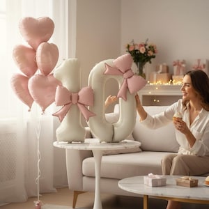 Peut inclure: Une scène festive avec des ballons en forme de cœur et des ballons numérotés formant le chiffre "10". Les ballons numérotés sont de couleur crème avec des nœuds roses. Une femme tient un cupcake.