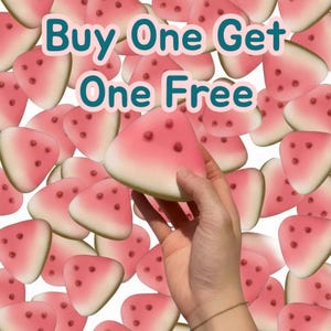 May include: A hand holding a watermelon-shaped item with a pink and white design and dark red dots. The image has the text "Buy One Get One Free" in a teal font with a pink outline. Many similar items are in the background.