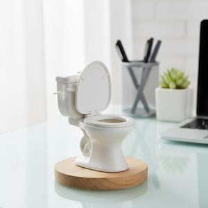May include: A miniature white toilet with the lid open, mounted on a round wooden base. The small ceramic toilet is a novelty item, likely a desk accessory or a decorative piece. The background includes a desk, a pen holder, and a small potted plant.