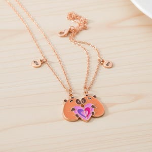 May include: Rose gold BFF necklaces. Each necklace features a pendant with two cartoon bear figures hugging a heart. The heart is divided into purple and pink sections. Small round charms with "BFF" are also included.