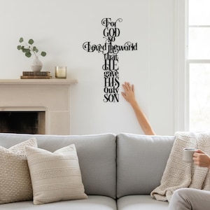 May include: Black metal wall art in the shape of a cross, featuring the words "For God so Loved the world that He gave His only Son." The art is mounted on a white wall. A person's arm is reaching towards the cross.