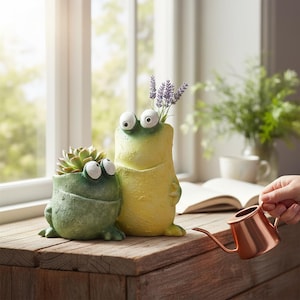 Puede incluir: Dos macetas con forma de rana, una verde y otra amarilla, están una al lado de la otra sobre una superficie de madera. La maceta verde contiene una suculenta, mientras que la amarilla tiene flores moradas. Una regadera de cobre está en primer plano.