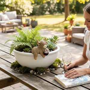 Puede incluir: Una maceta de cerámica blanca llena de plantas verdes y suculentas, acentuada con una figura de vaca de las Tierras Altas marrón que sostiene una copa de vino. La maceta está sobre una mesa de madera, rodeada de musgo y piedras pequeñas.