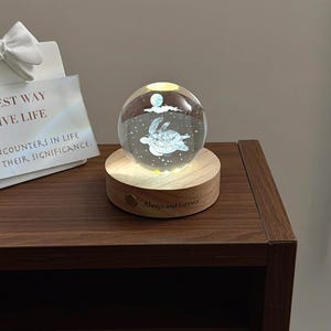 May include: A clear glass sphere featuring an internal laser-etched sea turtle and moon design, resting on a circular light-up wooden base with the engraved text Always and Forever. A white sign with a bow displays the text THE BEST WAY TO LIVE LIFE.