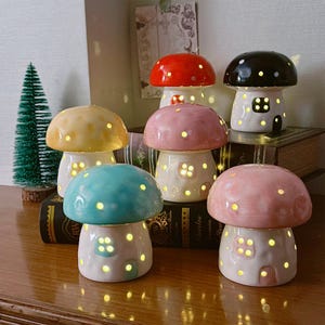 Può includere: Una collezione di sei lampade in ceramica a forma di fungo in vari colori, tra cui rosso, nero, giallo, blu e rosa. Ogni lampada presenta uno stelo cavo con finestre ritagliate e un cappello che brilla dall'interno, emettendo una luce calda.