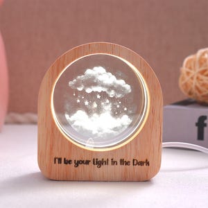 May include: A wooden night light with a clear, circular center displaying a cloud and star design. The light is framed by a wooden arch and features the text "I'll be your Light in the Dark" at the bottom. A white cord extends from the base.