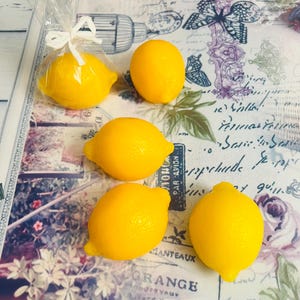 Può includere: Diversi limoni giallo brillante, alcuni singolarmente e altri avvolti in cellophane con un fiocco bianco, sono disposti su una superficie a motivi. I limoni hanno un aspetto strutturato e un colore vivace.