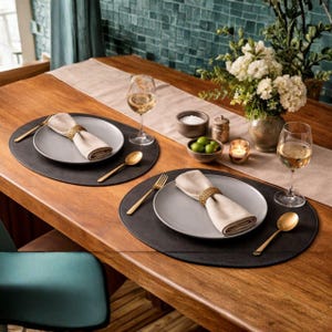 May include: A dining table set for two with gray plates, gold cutlery, and black placemats. Each place setting includes a folded napkin secured with a woven ring. Wine glasses and a floral arrangement complete the elegant setup.
