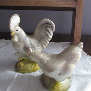 May include: A white ceramic rooster and hen salt and pepper shaker set. The rooster has a yellow base and a raised tail. The hen has a yellow base and is facing the left. The set is on a white lace tablecloth.