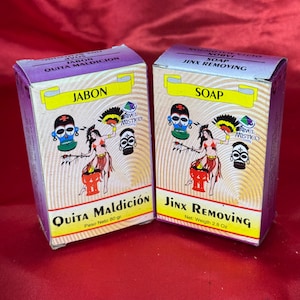 Op de afbeelding: Twee zeepdozen met kleurrijke afbeeldingen. Op de ene doos staat "JABON QUITA MALDICION" en op de andere "SOAP JINX REMOVING". Elke doos heeft een gestileerde illustratie van een vrouw en een schedel.