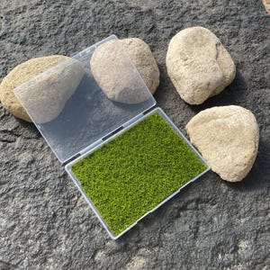 May include: A clear plastic container filled with vibrant green artificial grass, partially open, sits on a dark gray stone surface. Several light brown stones are scattered around the container, creating a naturalistic scene.