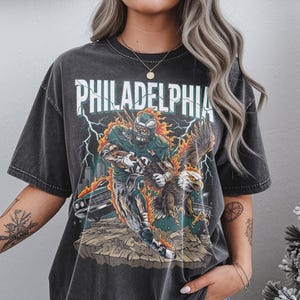 May include: Dark grey t-shirt with a distressed graphic. The design features the word "PHILADELPHIA" above an illustration of a football player and an eagle, with lightning bolts and a cityscape.