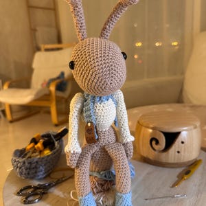 Puede incluir: Muñeca de ganchillo hecha a mano con forma de insecto, con ojos grandes y antenas. La muñeca es principalmente de color beige con detalles blancos y azules, con una bufanda y botas. Una pequeña etiqueta con las iniciales "C&K" está adjunta.