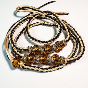 Puede incluir: Pulseras de cordón trenzado con cuentas color ámbar. Las pulseras presentan una combinación de cordones marrones, beige y dorados. Las cuentas son de color ámbar cálido con detalles plateados. Las pulseras se muestran sobre un fondo blanco.