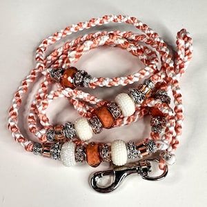 Peut inclure: Laisse pour chien tressée blanche et corail avec des perles décoratives blanches, orange et argentées. La laisse est dotée d'un mousqueton en métal argenté. Les perles sont rehaussées de métal argenté et or rose.
