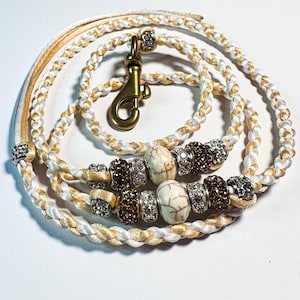 Może przedstawiać: Pleciona smycz dla psa w kolorze kremowym, białym i złotym. Smycz posiada ozdobne koraliki w kolorze brązowym, białym i srebrnym, ze złotym zapięciem. Przeznaczona dla zwierząt.