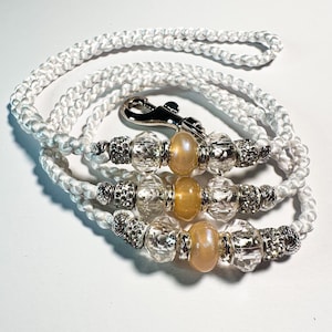 May include: White braided lanyard with decorative beads. The lanyard features clear, faceted beads, silver-toned accents, and peach-colored beads. A silver-toned clasp is at the top. The lanyard is designed for keys or an ID badge.