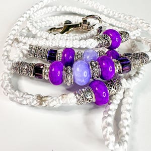 Peut inclure: Laisse blanche tressée pour chien avec des perles décoratives violettes et des accents argentés. La laisse est ornée d'un fermoir argenté. Les perles sont un mélange de violet vif et clair.