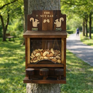 May include: A dark brown wooden squirrel feeder with a clear front panel filled with mixed nuts. The top section features the text THE NUT BAR and two light-colored squirrel silhouettes. Three small wooden pegs are attached to the bottom front.