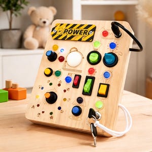 Puede incluir: Tablero de actividades de madera con varios botones, interruptores y perillas de diferentes colores. El tablero incluye la palabra "POWER" en un diseño de rayas amarillas y negras. Un juego de llaves está adjunto al tablero.