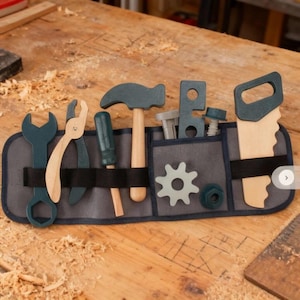 Puede incluir: Un juego de herramientas de juguete de madera con un cinturón de herramientas de tela gris y varias herramientas. Las herramientas incluyen una llave inglesa, unos alicates, un destornillador, un martillo, una sierra, un engranaje y tuercas y tornillos. Las herramientas están pintadas en tonos azules y de madera natural.