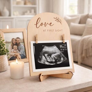 Puede incluir: Cartel de anuncio de embarazo de madera con las palabras "love at first sight" y una foto de ultrasonido. El cartel se exhibe sobre una mesa con una foto familiar enmarcada y una vela encendida.