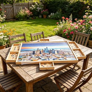 May include: A light-coloured wooden jigsaw puzzle table featuring a large central board with a completed city skyline puzzle and four pull-out drawers on the sides, each containing loose puzzle pieces, resting on a slatted wooden outdoor table.