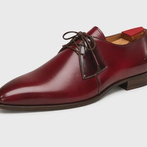 May include: A polished burgundy leather derby shoe with a smooth finish, dark brown laces, and a low stacked heel. The shoe features a subtle decorative perforation detail on the side heel area and includes a wooden shoe tree with a red top insert.