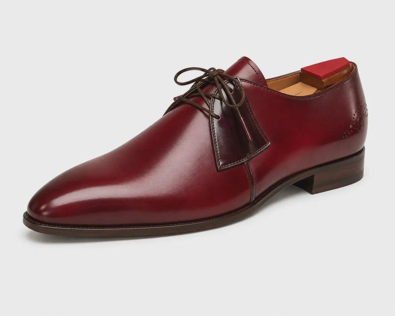 May include: A polished burgundy leather derby shoe with a smooth finish, dark brown laces, and a low stacked heel. The shoe features a subtle decorative perforation detail on the side heel area and includes a wooden shoe tree with a red top insert.