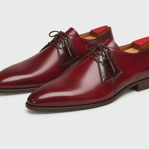 May include: A pair of polished burgundy leather derby shoes featuring a smooth finish, dark brown laces, and a low stacked heel. Each shoe includes a red wooden shoe tree inserted inside, visible at the heel opening, and a subtle embossed logo.