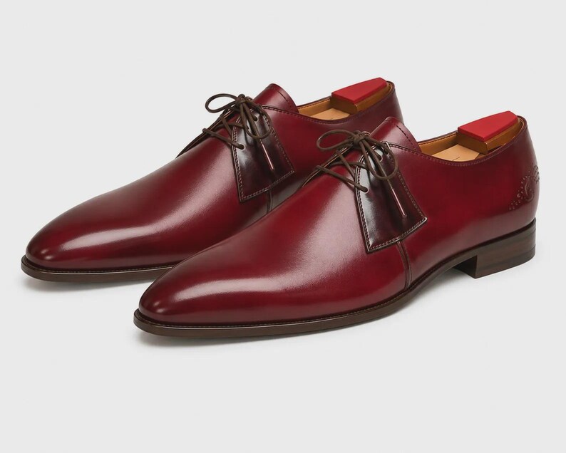 May include: A pair of polished burgundy leather derby shoes featuring a smooth finish, dark brown laces, and a low stacked heel. Each shoe includes a red wooden shoe tree inserted inside, visible at the heel opening, and a subtle embossed logo.