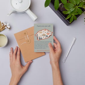 May include: A rectangular card with a gray background features the text Thank your past self at the top, followed by a central illustration of a hanging plant and botanical motifs, and a paragraph of text ending with And treat yourself with grace.