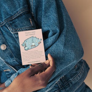 May include: A pink card with the text "Thank a taste you enjoyed" and a cartoon illustration of a blue creature with a cup. The card is held against a denim jacket.