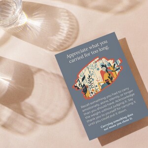 May include: A grey greeting card with the text "Appreciate what you carried for too long." The card features an illustration of plants and people. The card is on a light brown surface with two glasses.