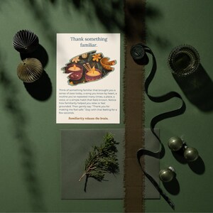 May include: An overhead shot features a greeting card with the text "Thank something familiar." Decorative paper ornaments, a sprig of greenery, and a dark ribbon are arranged on a green surface. The card's text is visible.