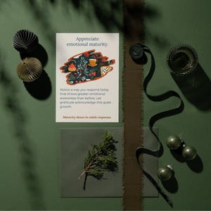 May include: A still life on a green surface includes a card with the text "Appreciate emotional maturity." and an illustration. Decorative paper balls, a sprig of greenery, and ornaments are arranged with a dark ribbon and a glass container.