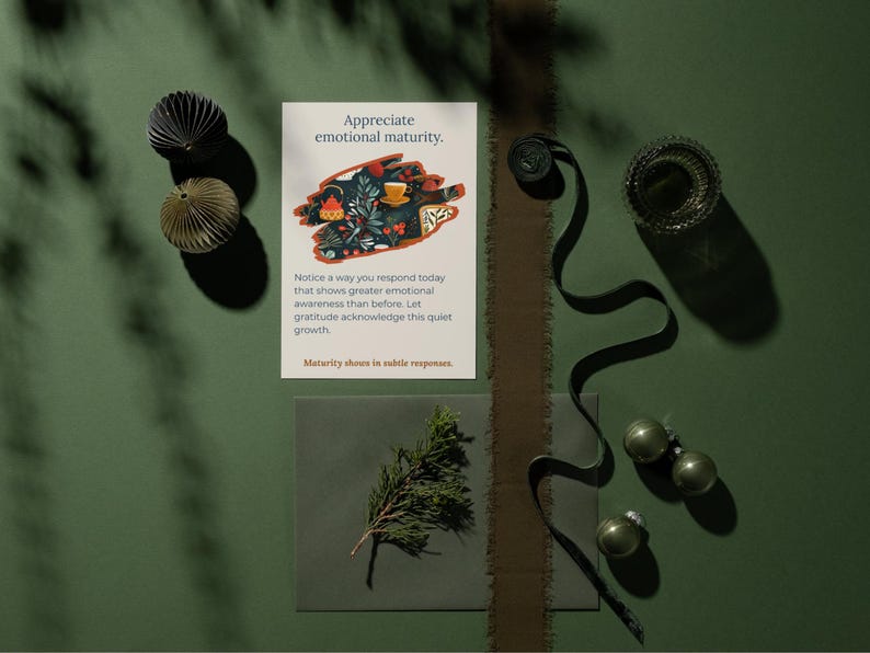 May include: A still life on a green surface includes a card with the text "Appreciate emotional maturity." and an illustration. Decorative paper balls, a sprig of greenery, and ornaments are arranged with a dark ribbon and a glass container.