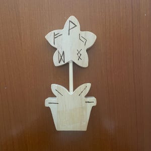 Peut inclure: Décoration en bois en forme de fleur, avec un sommet en forme d'étoile et une base en forme de pot. L'étoile porte des runes gravées. La fleur est reliée au pot par une tige en bois. La décoration est en bois clair.