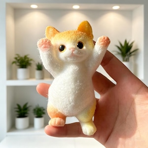 May include: A handcrafted cat ornament in white and orange, with its arms raised. The cat features large, dark eyes and pink paws. The figurine is made from a soft, textured material, possibly felt or wool. The cat's ears and head are orange.