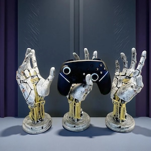 以下が含まれることがあります： 銀、金、白のロボットハンドの彫刻が3つ、黒いビデオゲームコントローラーを持っています。 手は機械的な要素で詳細に表現され、丸い台座に立っています。 コントローラーには白い照明アクセントがあります。