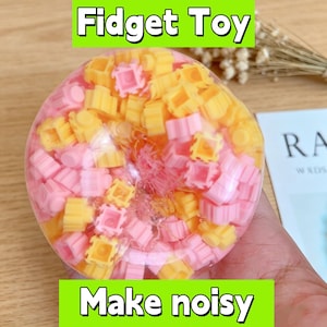 Op de afbeelding: Een doorzichtig, rond fidget-speeltje gevuld met kleine, kleurrijke plastic stukjes. Het speelgoed bevat gele en roze blokken, met de tekst "Fidget Toy" en "Make noisy" in groene vakken.