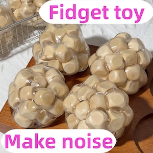 Puede incluir: Cuatro juguetes fidget de madera beige, cada uno compuesto por numerosos cubos pequeños, se muestran en bolsas de plástico transparente. El texto rosa "Fidget toy" y "Make noise" añade un toque lúdico.