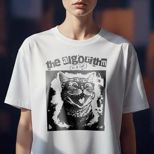 Op de afbeelding: Wit T-shirt met een zwart-witte afbeelding van een grijnzende kat en de tekst "the algorithm is a cat". De top heeft een ronde hals.