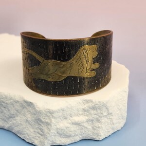 May include: A wide cuff bracelet made of dark, oxidised metal featuring a textured surface with small vertical indentations. A polished gold-tone metal inlay of a leaping lion is centred on the front of the band, which has an open back design.