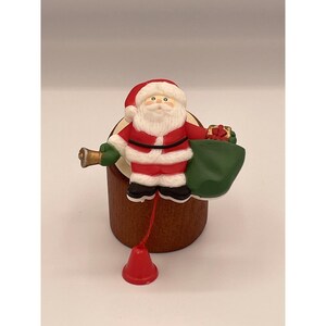 May include: A Christmas-themed music box featuring a Santa Claus figurine in a red suit with a white beard and hat. Santa holds a green sack and a small gift. A gold bell and a red bell are attached.
