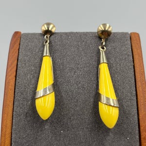 May include: A pair of yellow teardrop earrings with a silver band and gold-colored accents. The earrings have a post-back closure and are displayed on a gray surface.