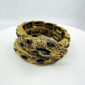 May include: A gold-toned metal cuff bracelet featuring a textured, scale-like surface. The bracelet is adorned with multiple teardrop-shaped black stones and small, clear, round crystals embedded along the center of the design.