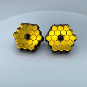 Puede incluir: Dos pendientes dorados con forma hexagonal y borde negro. Los pendientes presentan un diseño de panal y un pequeño centro oscuro. Se muestran sobre una superficie blanca.
